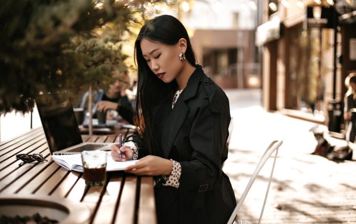 Charming young brunette lady in black trench coat make notes in notebook and works in laptop Asian