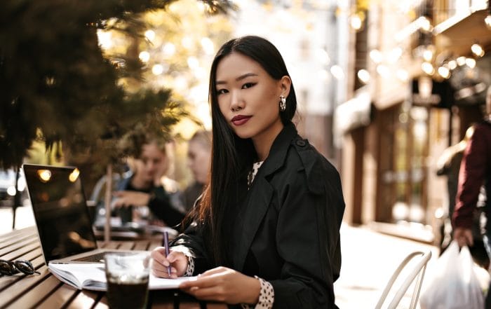 Beautiful young brunette Asian woman in black trench coat looks into camera sits at wooden desk ou